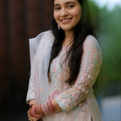 Woman in an embroidered pastel pink ethnic suit with floral patterns, matching net dupatta, henna-adorned hands, and pink bangles, standing outdoors against a soft, natural background.