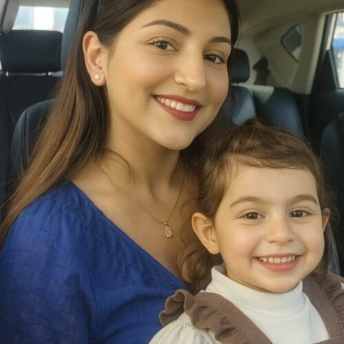 Woman with long straight brown hair, wearing a royal blue blouse and pearl stud earrings, sitting in a car with a young child on her lap. She has a delicate gold pendant necklace and sunglasses resting on her head. The child is dressed in a white top with a brown ruffled pinafore.