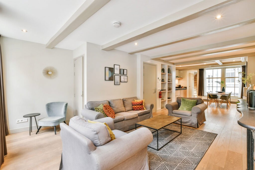 Open-plan modern living room with light gray sofas, matching armchairs with colorful accent pillows, wood flooring, minimalist coffee table and area rug, built-in shelves, neutral walls with framed art, stylish lighting, and a dining area near large windows.