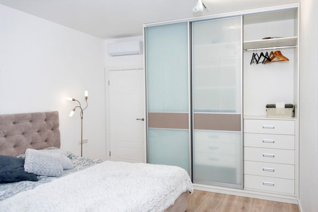 Minimalist sliding-door wardrobe with frosted glass panels, integrated drawers, open shelves, and hanging section in a bright, modern bedroom.